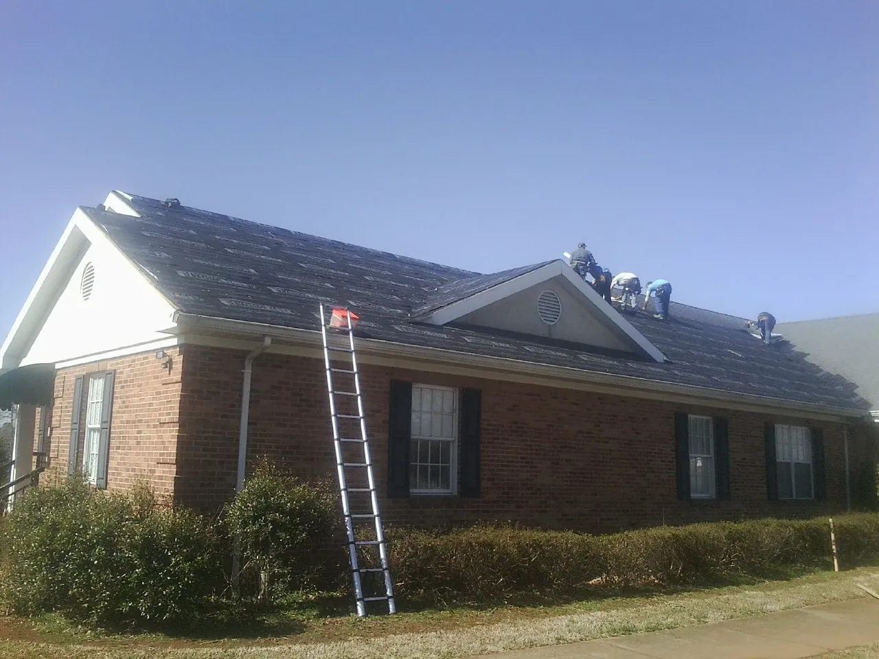 Crew tearing shingles off a brick ranch roof with a ladder set — Metal Roof Repair work by our crew in Surry