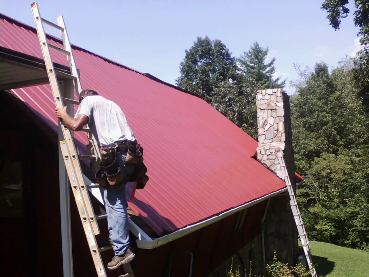 Roofer on a ladder beside a red metal roof and stone chimney — Soffit & Fascia Repair by Roofing Brothers in Surry